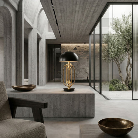 A modern minimalist interior with concrete walls and floors features the Ascot 4 Light Large Table Lamp in black & gold. Glass doors open to a courtyard tree, while bronze bowls and contemporary furniture complete the scene.