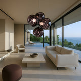 A modern living room features minimalist cream furniture, large glass windows with ocean and palm views, and a distinctive Lava LED 4 Light Chandelier - Smoked hanging from the ceiling.
