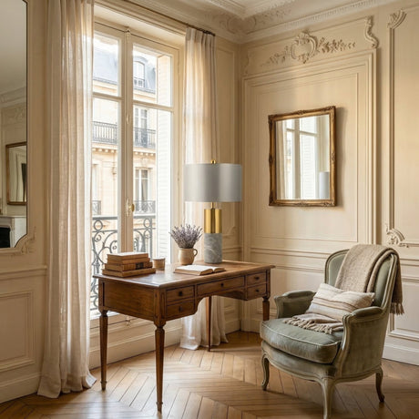 A sunlit elegant room with tall windows, a wooden desk topped with the Valmora 1 Light Monolith Marble Table Lamp in Grey & Brass and books, an upholstered armchair with throw blanket, wall mirror, herringbone wood floors, and ornate wall molding.