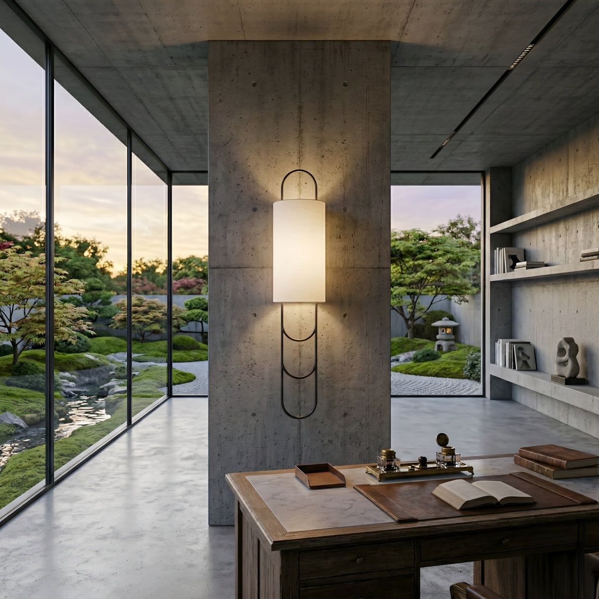 A modern office with a concrete pillar features the Catenis 2 Light Small Loop Frame Alabaster Wall Light in matte black and soft white, floor-to-ceiling garden-view windows, and a wooden desk with books and stationery in front.