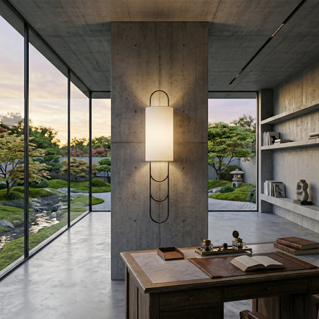 A modern office with a concrete pillar features the Catenis 2 Light Small Loop Frame Alabaster Wall Light in matte black and soft white, floor-to-ceiling garden-view windows, and a wooden desk with books and stationery in front.