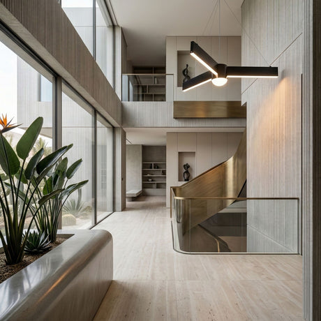 Modern interior with large windows, light stone floors, a curved metallic staircase, indoor plants on the left, minimalist furniture, and sculptural décor. The Fritz LED Suspension Light - Black hangs strikingly from the high ceiling.