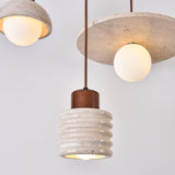 Rivano Ribbed Travertine Pendant Light in light beige stone with walnut-brown wood top, shown alongside a dome and disc pendant, each with glowing globe bulbs against a white background.
