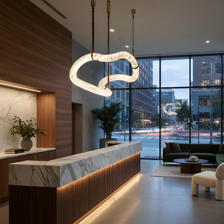 A modern hotel lobby features a marble reception desk, wood and marble accents, green plants, seating area, large floor-to-ceiling windows overlooking a city street at dusk, and the Serava LED Large Alabaster Asymmetric Suspension Light – Brown & Soft White.