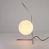 The Vetra 1 Light Opal Glass Table Lamp - Grey features a chrome metal frame with a diagonal rod, a glowing white opal glass shade, and a black power cord, displayed on a plain white surface.