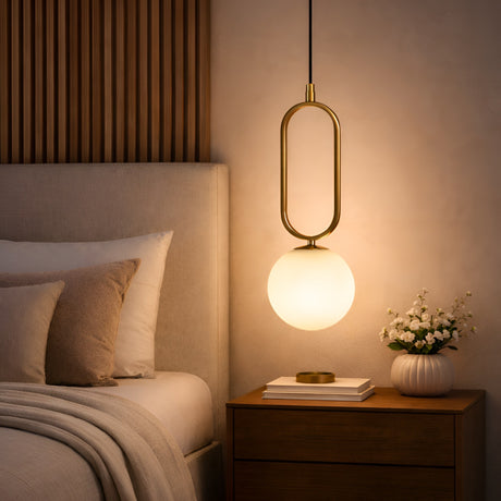 A modern bedroom with the Orvyn 1 Light Small Globe Alabaster Pendant in soft white and brushed brass hanging over a wooden nightstand, which holds books, a small gold dish, and a vase of white flowers beside a neatly made bed.