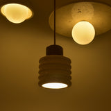 Rivano Ribbed Travertine Pendant Light in light beige stone with a walnut-brown wooden top, shown lit and hanging near two globe pendants on a textured ceiling.