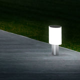 Tiny Round Pillar Light IP45 in satin nickel, illuminated white cylindrical shade on a slim metal stem, shown outdoors on a paved path edge at night.