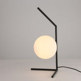 The Vetra 1 Light Opal Glass Table Lamp in black features a minimalist metal frame, glowing opal glass shade, and a black power cord, all set against a simple light background.