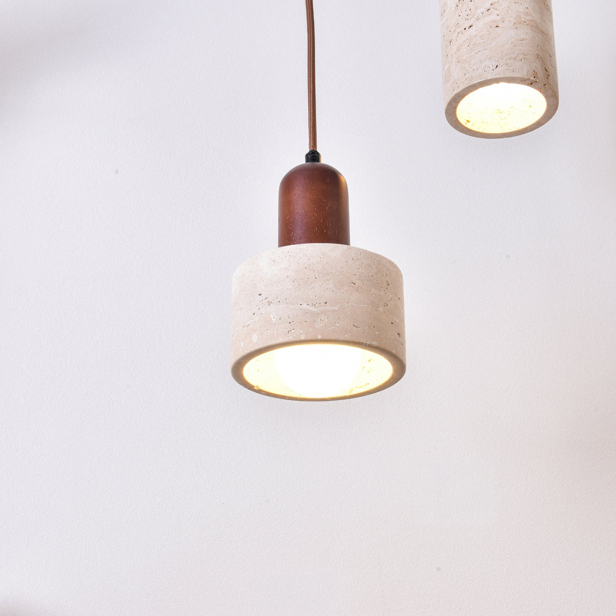 Torvella Travertine Drum Wall Light in light beige travertine with a walnut brown fitting, suspended on a fabric flex with a lit E27 bulb, shown against a plain white background.