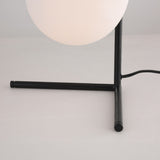 A close-up of the Vetra 1 Light Opal Glass Table Lamp in black, featuring a round opal glass shade and black metal tripod base on a light surface, with a power cord extending from the base.