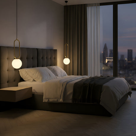 A modern bedroom at dusk with a large bed, tufted headboard, sheer curtains, and floor-to-ceiling windows overlooking the city, softly lit by the Orvyn 1 Light Small Globe Alabaster Pendant in Soft White & Brushed Brass.