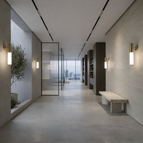 A modern, minimalist hallway with concrete floors, a Magari Wall Light - Gold, a stone bench, built-in shelves, and large windows at the end revealing a city view. An indoor tree stands to the left.