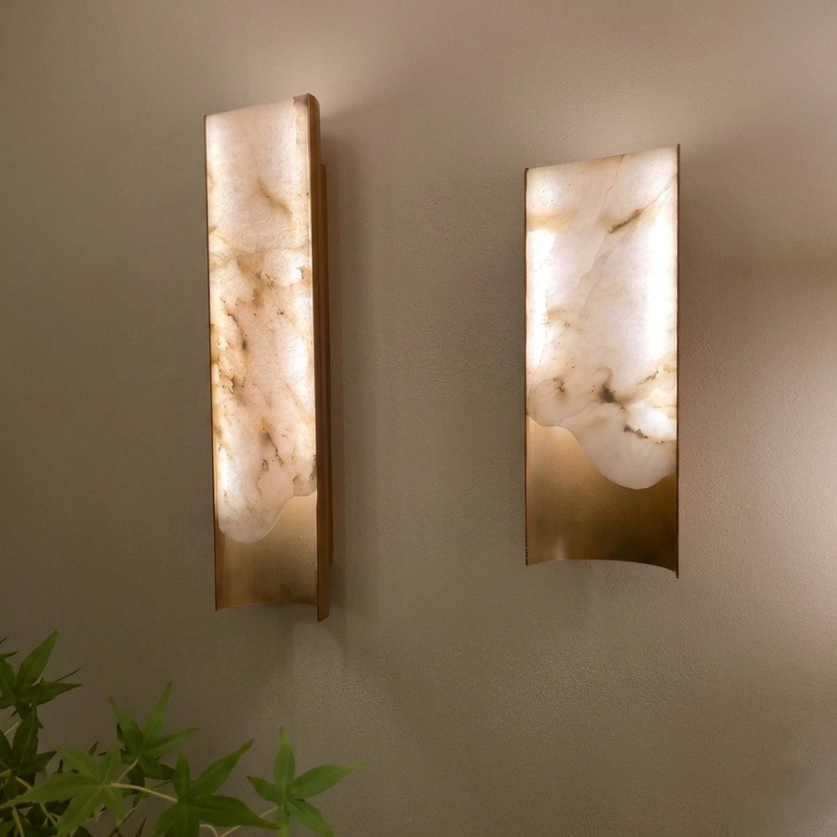 Pair of Caelith Medium wall lights with carved alabaster panels in brushed brass frames, glowing warmly on a neutral wall above a green plant.