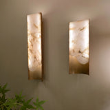 Caelith Small carved alabaster wall lights in brushed brass, curved vertical panels with rough-cut stone glowing softly, mounted on a neutral wall.