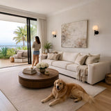 A woman stands by open glass doors with a golden retriever on a cream rug in a bright modern living room featuring neutral decor, a round wooden coffee table, and the Olivaro 1 Light Pedal Form Alabaster Wall Light. Palm trees and ocean are visible outside.