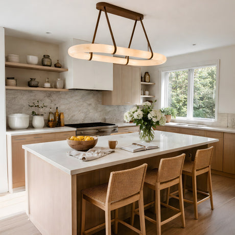 Modern kitchen with light wood cabinets, marble backsplash, and a large island with three woven barstools. The Cintura LED Small Alabaster Floating Suspension Light in soft white hangs above, highlighting white flowers and a bowl of lemons.