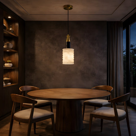 A round wooden dining table with four chairs sits beneath the Bellaro 1 Light Large Textured Column Alabaster Single Pendant Light – Soft White & Brass in a dim, elegant room with dark walls, shelves, and a curtained window.