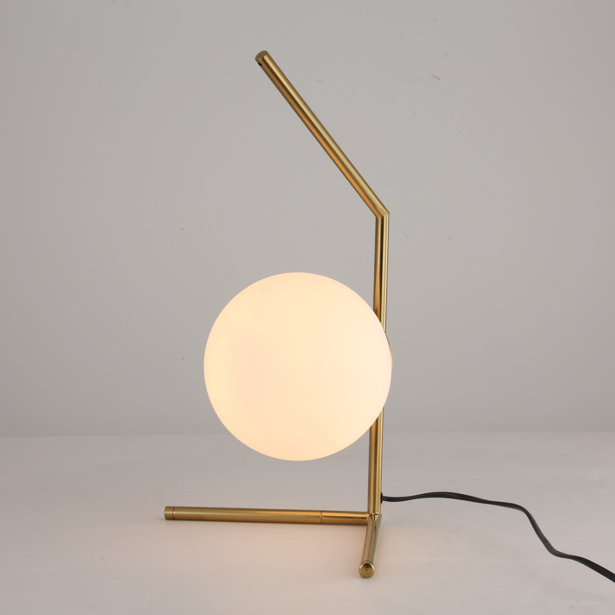 The Vetra 1 Light Opal Glass Table Lamp in gold features a metal frame and a frosted white opal globe shade, resting on a white surface with its black power cord visible on the right.