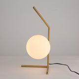 The Vetra 1 Light Opal Glass Table Lamp in gold features a metal frame and a frosted white opal globe shade, resting on a white surface with its black power cord visible on the right.
