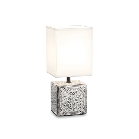 Product Description: The Glimmerlux Small Table Lamp - White features a tall, rectangular white shade and an ornate square ceramic base adorned with intricate silver patterns. This elegant piece beautifully fuses modern and classic styles, embodying sophisticated decorative lighting.