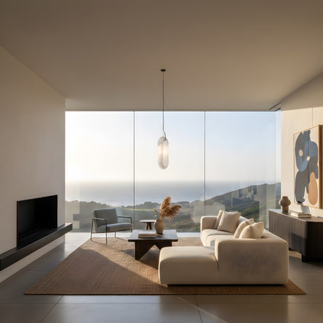 A modern living room features minimalist furniture, large ocean-view windows, a white sofa, armchair, abstract art, and a Serava LED Small Alabaster Single Pendant Light – Soft White above a central coffee table on a woven rug.