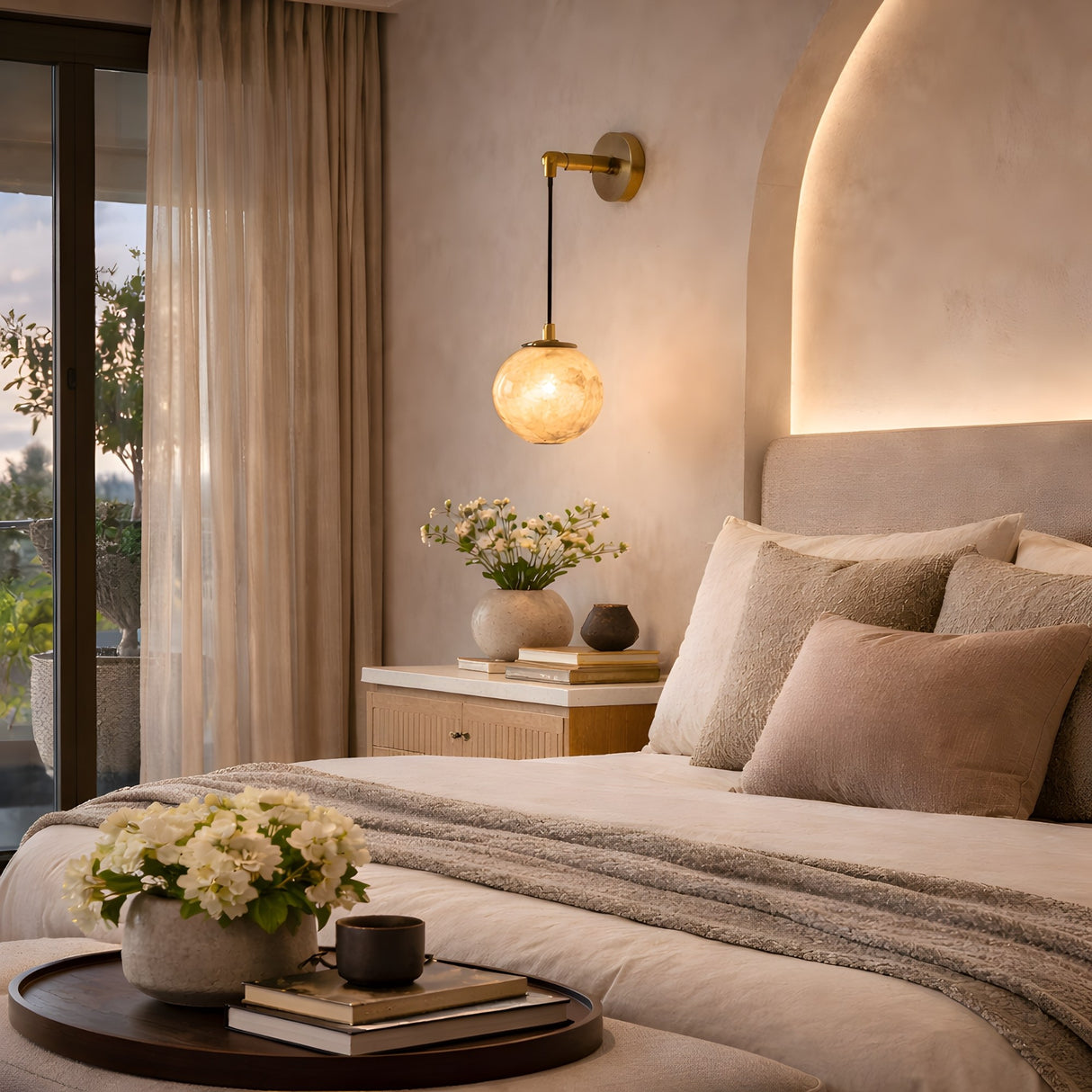 A cozy, modern bedroom with beige tones, soft bedding, a round tray with flowers and a mug on the bed, a nightstand with books and decor, warm lighting from the Drisa 1 Light Hanging Globe Alabaster Wall Light, and sheer curtains framing a balcony door.