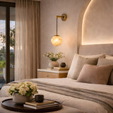 A cozy, modern bedroom with beige tones, soft bedding, a round tray with flowers and a mug on the bed, a nightstand with books and decor, warm lighting from the Drisa 1 Light Hanging Globe Alabaster Wall Light, and sheer curtains framing a balcony door.