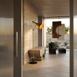 A modern, minimalist living room with concrete walls and floors, a Funk Floor Lamp - Brass, abstract wall art, and a black sofa by large windows overlooking a desert landscape.