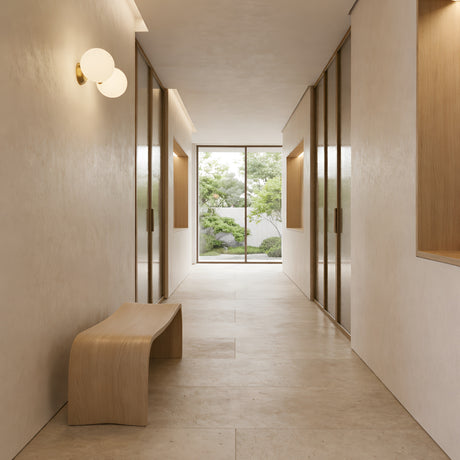 Minimalist hallway with light beige walls and floors, wooden accents, a curved wooden bench, the Vetra 2 Light Opal Glass Wall/Semi-Flush Ceiling Light in gold on the wall, and large glass doors opening to a lush garden.