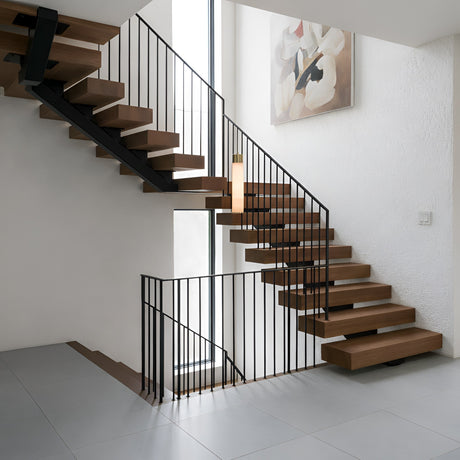 A modern wooden staircase with black metal railings stands in a bright, minimalist space. A tall window brightens the area, while the Norex LED Tube Alabaster Single Pendant Light in soft white and brushed brass hangs above an abstract painting.