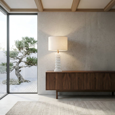 A modern, minimalist room features a wooden sideboard, the Echelon 1 Light Stepped Alabaster Table Lamp in brass and soft white, a large window, and views of a small sculpted tree in a gravel garden.