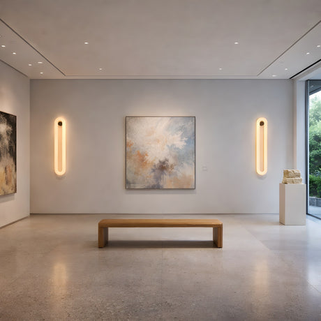 A modern art gallery with a wooden bench, abstract paintings, two Serava LED Large Alabaster Wall Lights – Soft White on the walls, and a sculpture by a window overlooking lush greenery.