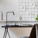 MiniSpot LED Table Lamp in black iron with round base, bent arm and cylindrical head, shown on a desk beside a mug, notebooks and a wooden mannequin.