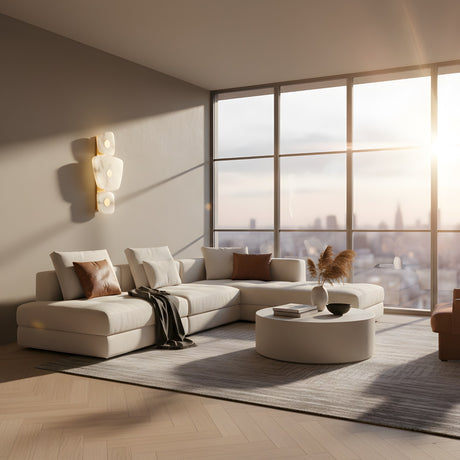 A modern living room with a large white sectional sofa, brown and white pillows, round coffee table, and floor-to-ceiling windows overlooks a sunset city skyline. The Calyra LED Alabaster Wall Light in Brass & Soft White adds elegance to the minimalist space.
