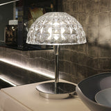 Deco Crystal Table Lamp in chrome with a faceted crystal dome shade on a slim stem and round base, lit on a shelf beside books and bowls.