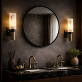 A modern bathroom featuring a dark marble double sink, brass faucets, a round mirror, Marvello 1 Light Cylinder Alabaster Wall Lights in soft white and matte black, plus decorative bottles, towels, and a potted plant on the countertop.