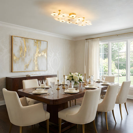 Elegant dining room featuring a dark wood table, eight white upholstered chairs, gold and white table settings, floral centerpiece, abstract gold artwork, large window with sheer curtains, and the Calyra LED Large Alabaster Semi-Flush Ceiling Light in Brass & Soft White.
