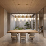A minimalist dining room features a wooden table, six chairs, the Magari 10 Linear Suspension Light in gold above, and large glass doors opening to a tranquil Japanese-style garden with rocks and a bonsai tree.