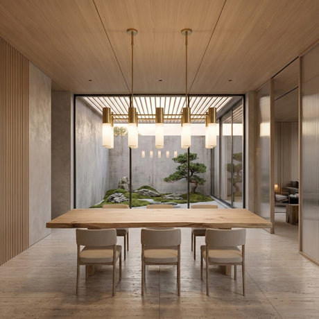 A minimalist dining room features a wooden table, six chairs, the Magari 10 Linear Suspension Light in gold above, and large glass doors opening to a tranquil Japanese-style garden with rocks and a bonsai tree.