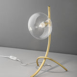 Dolce 1 Light Glass Table Lamp with a matt gold curved metal base and round clear glass shade, G9 bulb visible, on a plain grey background.