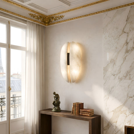 The Majorae LED Large Split Alabaster Wall Light in Soft White & Matte Black casts a gentle glow on a white wall near marble panels. A wooden console below holds books and a sculpture, with sheer curtains framing the Eiffel Tower view.
