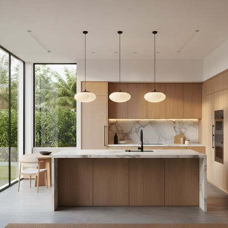 A modern kitchen with wood cabinets, marble island and backsplash, built-in appliances, and large windows overlooks greenery. Above the island hang three Solenne 1 Light Minimal Orb Alabaster Pendants in soft white & matte black; a dining table for two sits in the corner.