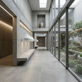 A modern minimalist hallway with concrete walls and floors, built-in benches, large glass windows overlooking an indoor garden with plants and a stone planter, and a Funk Stout Large Pendant Light - White hanging overhead.