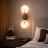 A modern bedroom with a beige upholstered headboard, wooden nightstand with a vase, books, and bowl, and the Althia LED Alabaster Wall Light in brushed brass & soft white adds decorative lighting with its two glowing lamps.