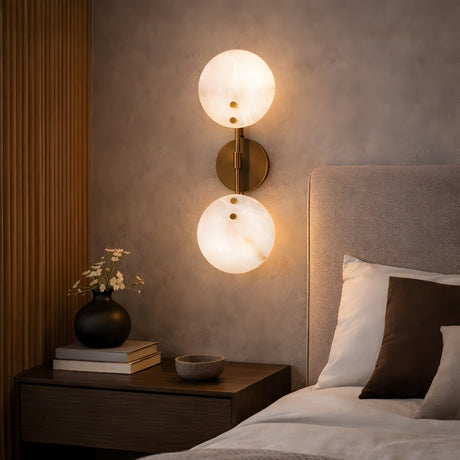 A modern bedroom with a beige upholstered headboard, wooden nightstand with a vase, books, and bowl, and the Althia LED Alabaster Wall Light in brushed brass & soft white adds decorative lighting with its two glowing lamps.