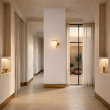 A minimalist hallway with beige walls and floors features built-in lighted shelves, a central wall with the Vetra 1 Light Opal Glass Wall Light in gold, and sheer curtains framing glass doors that open to a patio filled with potted plants.