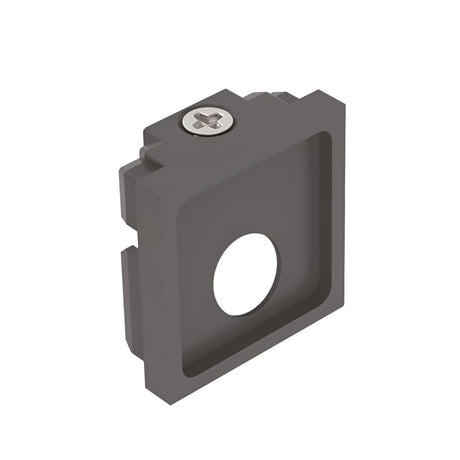 The Luxeon-1 Small End Power Cap for 3-Phase Track in black is a square metal bracket frequently utilized as an end cap in track lighting systems. It features a central circular hole and a protruding screw at the top, with several grooves and edges that give it a three-dimensional appearance. The bracket is shown isolated against a white background.