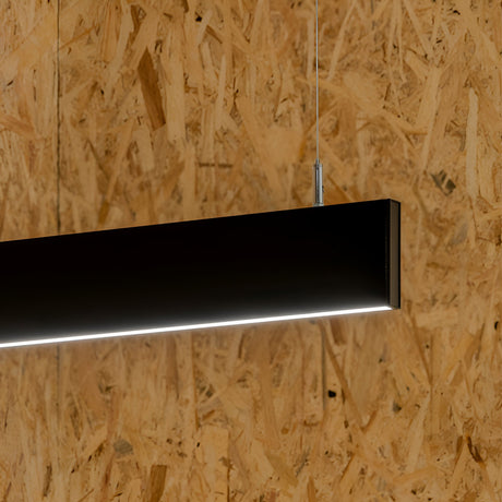 A sleek black Flux LED Linear Suspension 32W 4000K fixture hangs from the ceiling, offering a modern lighting solution that emits a soft white glow, beautifully contrasting with the textured wooden background. Additionally, it's dimmable for perfect ambiance control.