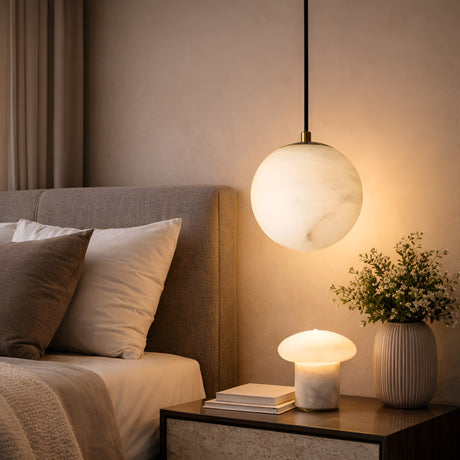 A cozy bedroom featuring a gray upholstered bed, white pillows, the Lyvane 1 Light Extra Small Globe Alabaster Single Pendant Light in soft white, a mushroom-shaped table lamp, and a vase of flowers on a nightstand against a beige wall.